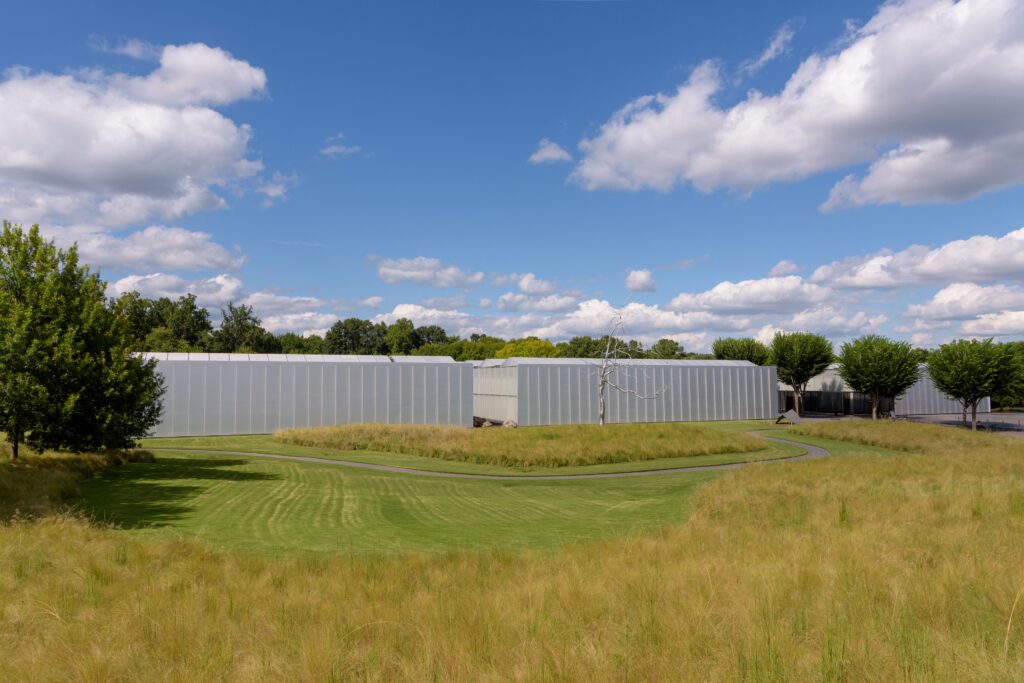 An aluminum and glass modernist building surrounded by manicured meadows and deciduous trees.