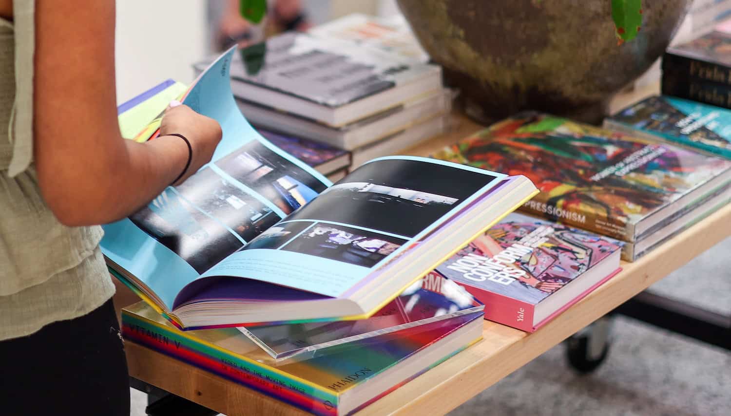 A person turns the page of a book, standing in front of a table covered in colorful books.