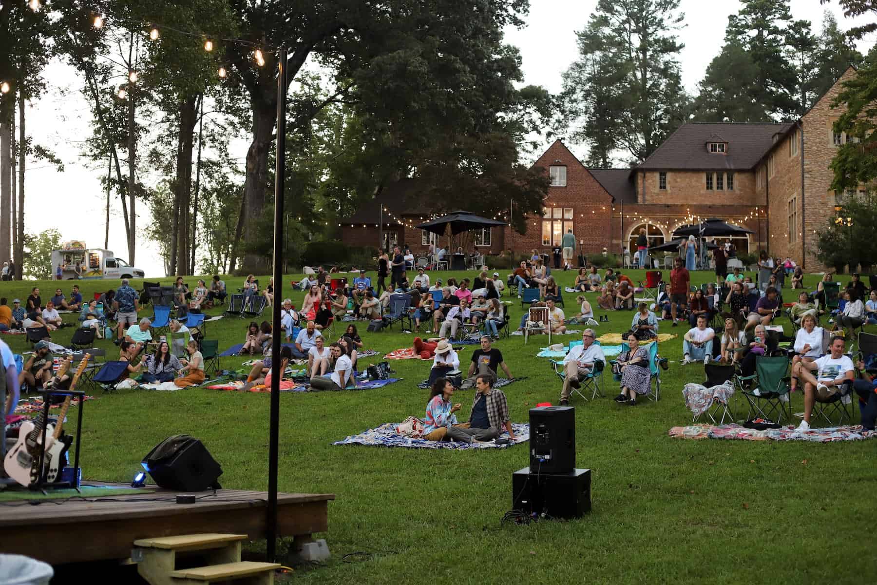 Concertgoers sit in lawn chairs and blankets spread out on a grassy hillside below a historic home.